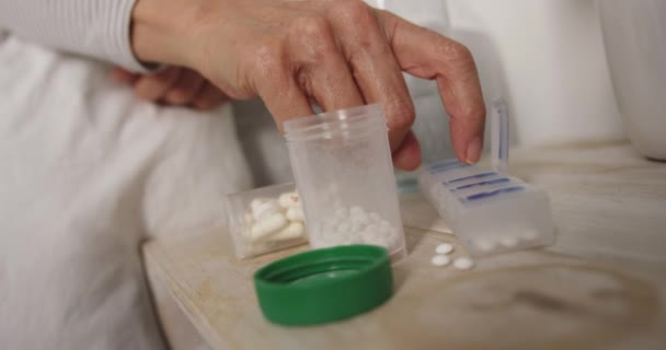 Close up of the hand of a senior Caucasian woman in her bed at home, reaching to her bedside table and taking a tablet out of a weekly pill box, with other containers of pills and a glass of water on the bedside table, slow motion - 映像、動画