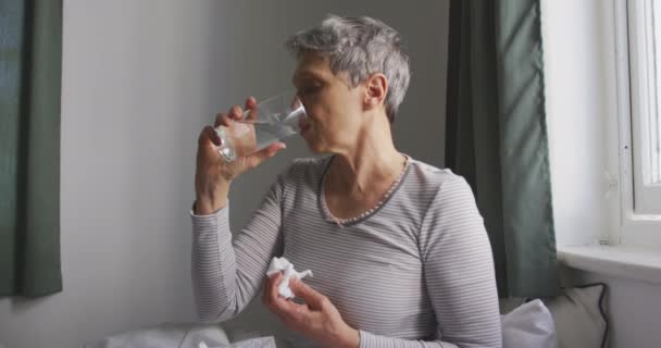 Side view of a senior Caucasian woman with short grey hair sitting on her bed and drinking a glass of water and holding a tissue in front of the window in her bedroom at home, slow motion - 映像、動画