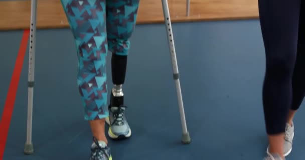 Front view of a Caucasian woman with a prosthetic leg walking on crutches at a sports centre gym with a Caucasian female physiotherapist guiding her through an exercise programme holding a clipboard - Filmagem, Vídeo