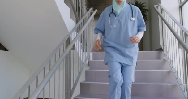 Front view of a mixed race female healthcare worker wearing scrubs, a stethoscope and a hijab running down the stairs in a hospital, slow motion - Video