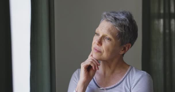 Side view close up of a senior Caucasian woman with short grey hair standing and looking out of the window at home with her hand under her chin, slow motion - Felvétel, videó