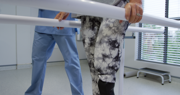 Side view of a Caucasian male physiotherapist helping a Caucasian female patient with a prosthetic leg walk between parallel bars during a physiotherapy session at a hospital, slow motion - Materiał filmowy, wideo