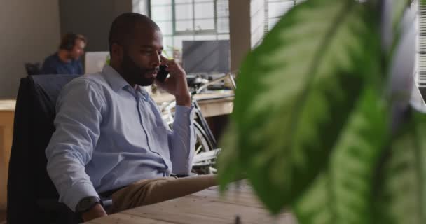 Vista lateral de un joven profesional creativo de raza mixta en una oficina moderna, trabajando en un escritorio hablando en un teléfono inteligente con su colega en segundo plano, cámara lenta
 - Imágenes, Vídeo