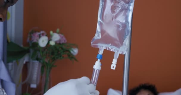 Side view of an African American male healthcare worker checking the IV bag and tubes hanging above the bed of a patient in a hospital ward, slow motion - Πλάνα, βίντεο