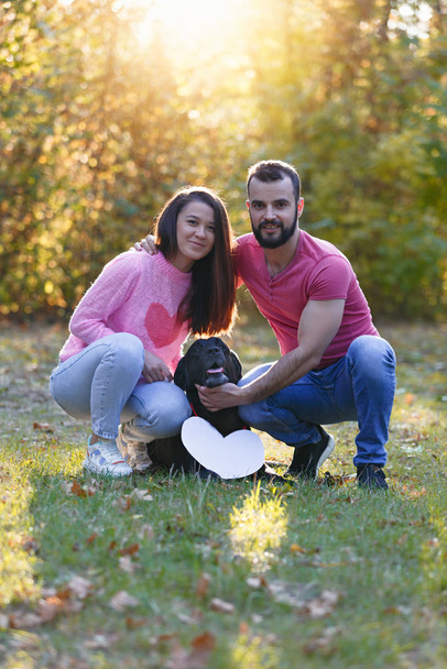 Mladý zamilovaný pár objímající se v parku. Mezi nimi je černý labrador s bílým znakem ve tvaru srdce. Koncept šťastného rodinného života v lásce. - Fotografie, Obrázek