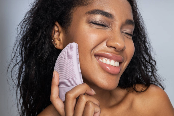 glimlachend Afro Amerikaans meisje houden siliconen borstel voor gezicht in haar hand - Foto, afbeelding