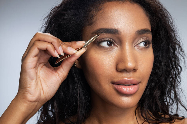 Afro American woman is plucking eyebrows with tweezers - Foto, afbeelding