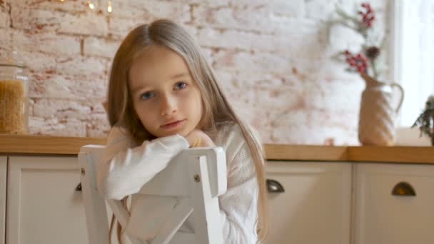 kleines trauriges Mädchen mit blauen Augen und langen blonden Haaren in weißem Kleid und Pullover sitzt auf einem Stuhl in einem Raum im Loft-Stil - Filmmaterial, Video