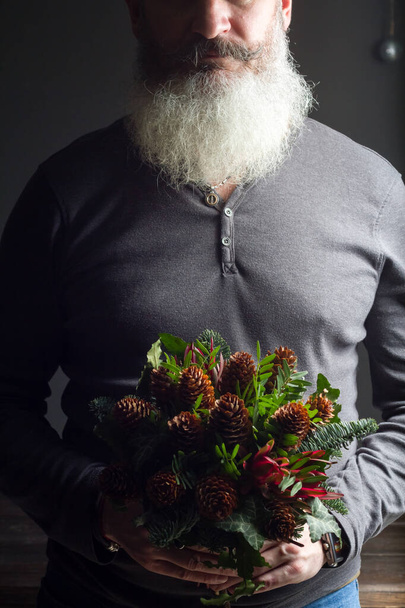 Uomo barbuto di mezza età in possesso di un bouquet invernale di ramoscelli di abete nobile, coni, pistacchio, leucodendro e edera, concetto regalo invernale
 - Foto, immagini