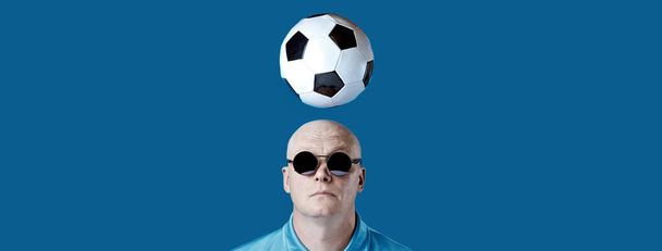 bald brutal man in dark round glasses with highlights. A football hovered overhead. On a blue background. - Φωτογραφία, εικόνα