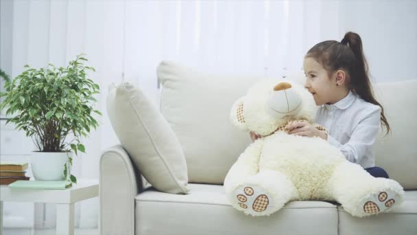 Cute little girl sitting on the sofa, talking with her teddy, whispering some secret in its ear. - Imágenes, Vídeo