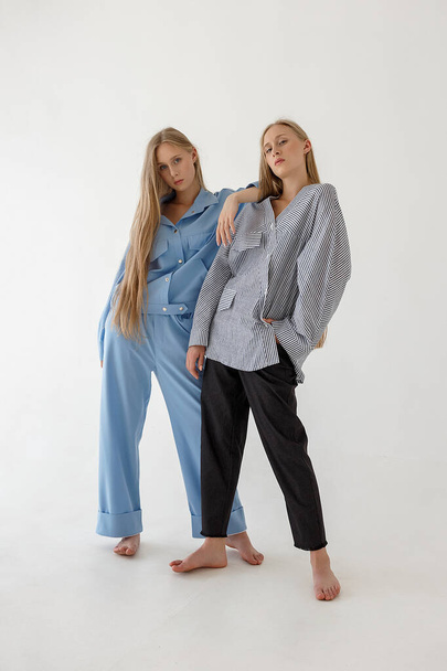 two pretty young twin sisters with long blond hair posing on white background in oversize clothes. Fashion photoshoot - 写真・画像