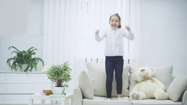 Cheerful little girl is bouncing on the sofa joyfully, smiling. Soft teddy near her. - Felvétel, videó