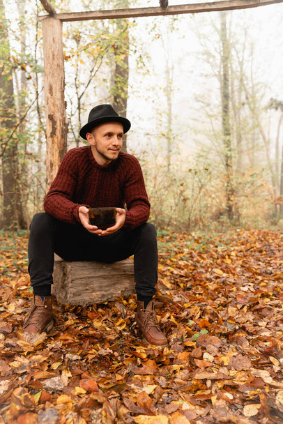 Hombre guapo feliz en sombrero negro y suéter rojo sentado en el muñón con taza de cofee, estar en el colorido bosque
 - Foto, Imagen