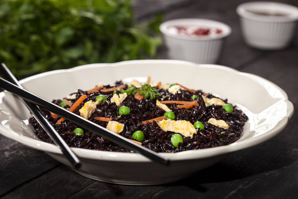 Cooked black rice with vegetables in a plate with sauces and chopsticks on the wooden background. - Photo, image