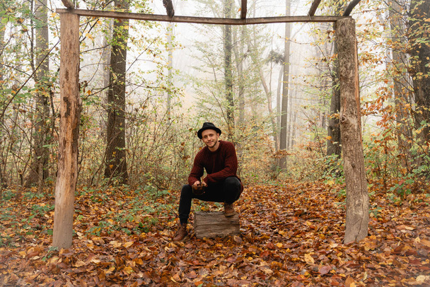 Handsome yound stylish man in black hat and red sweater crouched on the stump among the colorful forest - Valokuva, kuva