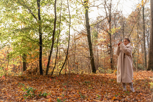 Menina bonita morena atraente em casaco branco andando entre a floresta colorida e fazendo foto em seu telefone celular
 - Foto, Imagem