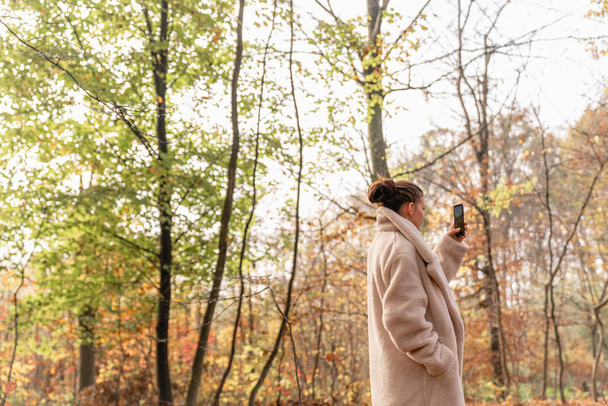 Приваблива красива брюнетка дівчина в білому пальто ходить серед барвистого лісу і робить фото на її мобільному телефоні
 - Фото, зображення