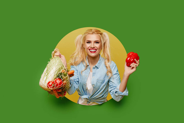 Молодая женщина собирала свежие овощи в пакете на зеленом фоне. Подглядывает из круглого отверстия в стене. Retro and pin up style. Концепция правильного питания и вегетарианства
. - Фото, изображение