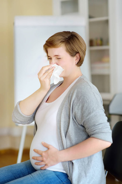 Hamile genç bir kadın hapşırıyor ve doktorun kabulü sırasında peçeteyle burnunu siliyor. Grip mevsimi, soğuk algınlığı. Alerjik insanlar.. - Fotoğraf, Görsel