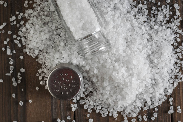 Μια ανοιχτή αλατιέρα σε μια στοίβα χυμένο αλάτι σε ένα ξύλινο τραπέζι. - Φωτογραφία, εικόνα