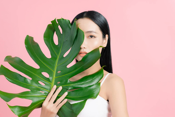 Portrait of young beautiful Asian woman holding tropical leaf on pink background. Natural beauty concept. - Foto, Imagem