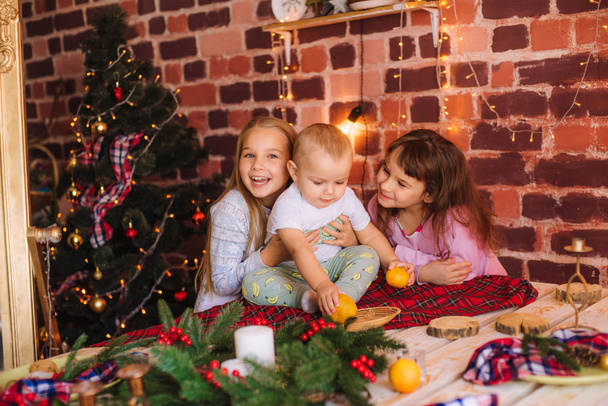 Cute sisters and little brother in pajamas have fun in the kitchen by the table with gingerbread cookies, tangerines at home in the room decorated with Christmas garlands, needles. . Christmas mood - Foto, Imagem
