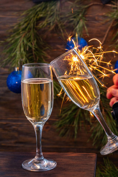 Glasses of champagne with bubbles on the background of Christmas decorations. Two glasses touch each other during a festive toast and sparklers burn beautifully. Beautiful card. - Фото, зображення