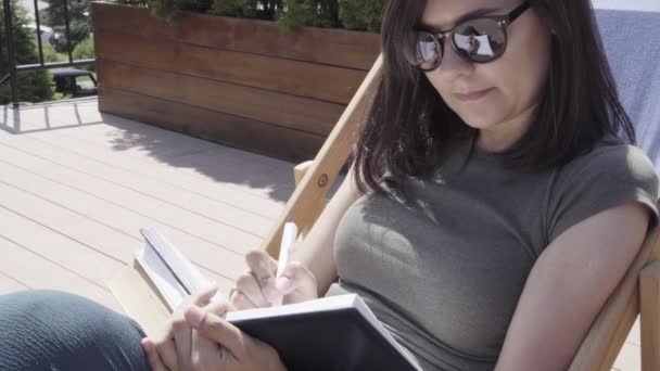 Young woman in sunglasses taking notes in notebook outdoors - 映像、動画
