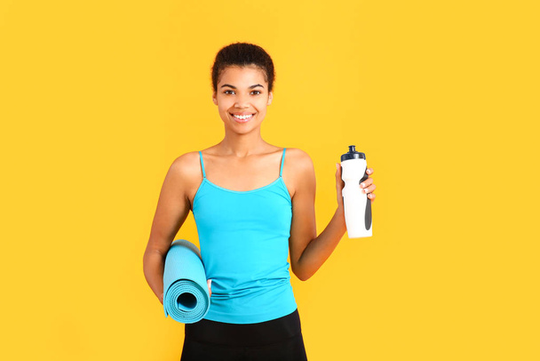 Sportovní Afroameričanka s lahví vody a jóga podložka na barevném pozadí - Fotografie, Obrázek