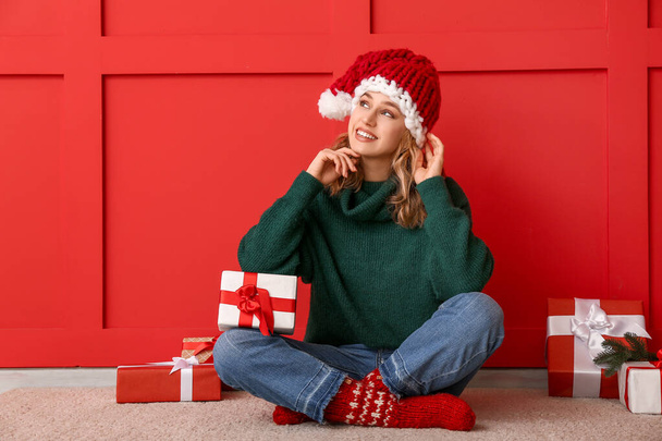 Happy young woman in winter clothes and with Christmas gifts sitting near color wall - Φωτογραφία, εικόνα