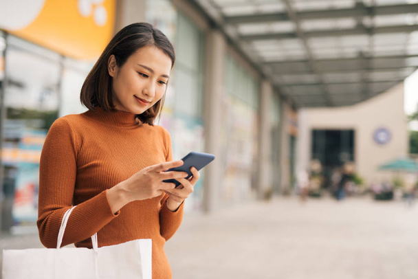 Portré elegáns fiatal ázsiai nő gazdaság bevásárló táskák és használata okostelefon útközben elhagyja bevásárlóközpont - Fotó, kép
