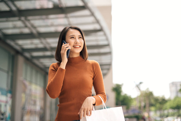 Portrait of happy young Asian woman talking on mobile and carrying paper shopping bags walking outdoor mall - Foto, Imagen
