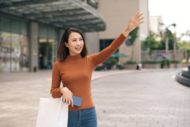 Alegre joven mujer asiática con bolsa de compras captura de taxi fuera del centro comercial
 - Foto, Imagen