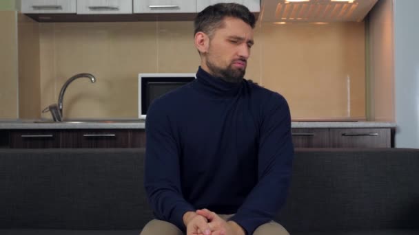 Een jongeman in een donkerblauwe trui en grijze broek zit op de bank en is scheef van wat hij zag op TV. Keuken op achtergrond. 4k slow motion beelden - Video