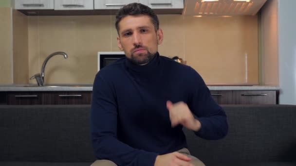 Ένας νεαρός άνδρας με σκούρο μπλε πουλόβερ και γκρι παντελόνι κάθεται στον καναπέ και θυμώνει και ορκίζεται σε μια τηλεοπτική εκπομπή. Κουζίνα στο παρασκήνιο. 4k βίντεο βραδείας κίνησης - Πλάνα, βίντεο
