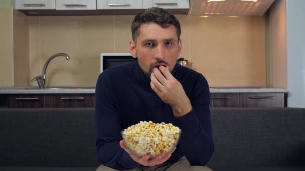 A young man in a dark blue pullover and gray pants is sitting on the couch, eats popcorn from a transparent glass plate and watches entertainment on TV. Kitchen on background. 4k slowmotion footage - Metraje, vídeo