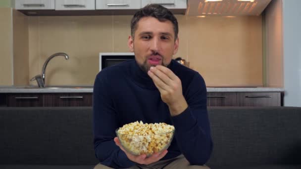 A young man in dark blue pullover and gray pants sitting on the couch, eats popcorn from transparent glass plate, smiles and watches entertainment on TV. Kitchen on background. 4k slowmotion footage - Metraje, vídeo