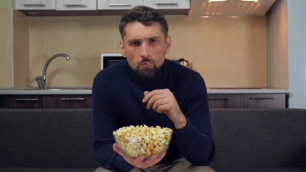Serious young man in dark blue pullover and gray pants sitting on the couch, eats popcorn from transparent glass plate and gets angry, swears on a TV show. Kitchen on background. 4k slowmotion footage - Séquence, vidéo