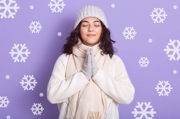 Portrait of pretty lady, wearing cap, mittens, sweater, standing and praying, keeps hands together in front of chest, posing with closed eyes, lady having dark wavy hair, poses against studio wall. - Foto, Bild