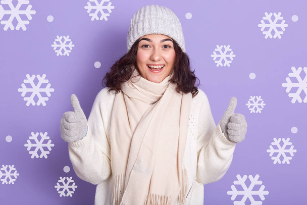 Winter girl with dark wavy hair wearing white warm sweater, scarf, cap and mittens standing with thumbs up isolated over studio wall with snowflake on background, girl looks happy and excited. - Photo, Image