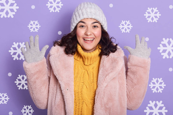 Image of woman wearing stylish eco fur coat, yellow sweater and cap, looking at camera and keeping hands up in studio with snowflakes on background. Female looks excited and expresses happyness. - Foto, imagen