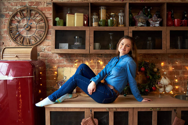 Pretty woman in jeans clothing sitting on table in kitchen at home with Christmas decor and garland on background - Foto, afbeelding