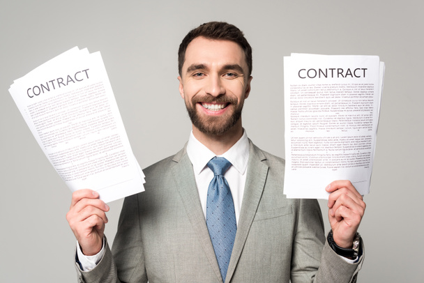 щасливий бізнесмен тримає контракти і дивиться на камеру ізольовано на сірому
 - Фото, зображення