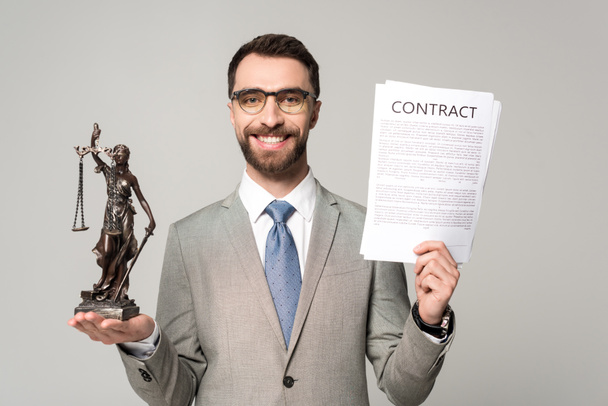 handsome lawyer holding contract and themis statue while smiling at camera isolated on grey - Photo, Image
