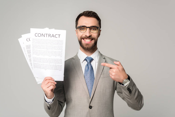 hombre de negocios sonriente en gafas apuntando con el dedo a los contratos aislados en gris
 - Foto, imagen