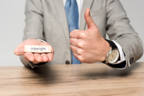καλλιεργημένη άποψη του επιχειρηματία που κρατά ξύλινο μπλοκ με λέξη copyright και δείχνει τον αντίχειρα προς τα επάνω απομονώνονται σε γκρι - Φωτογραφία, εικόνα