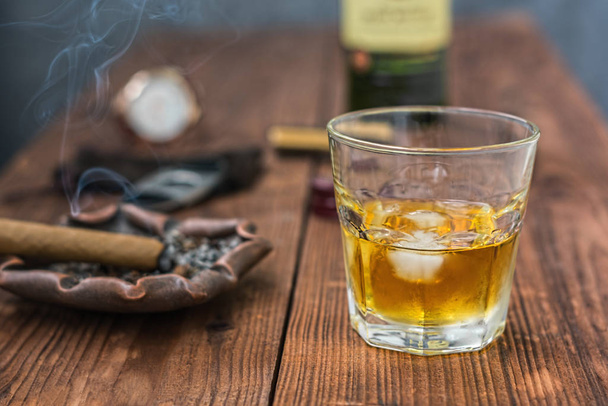 ein Glas Whisky mit Eis auf einem Tisch im Vordergrund und eine Zigarre mit einem Aschenbecher, einer Guillotine, einem Feuerzeug, einem Fragment einer Whiskeyflasche und einer Uhr im Hintergrund in Unschärfe - Foto, Bild