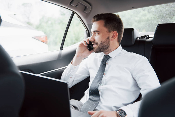 A young businessman working on laptop and talking on the phone while sitting in the car's back. Works in motion, appreciates its time. - Valokuva, kuva