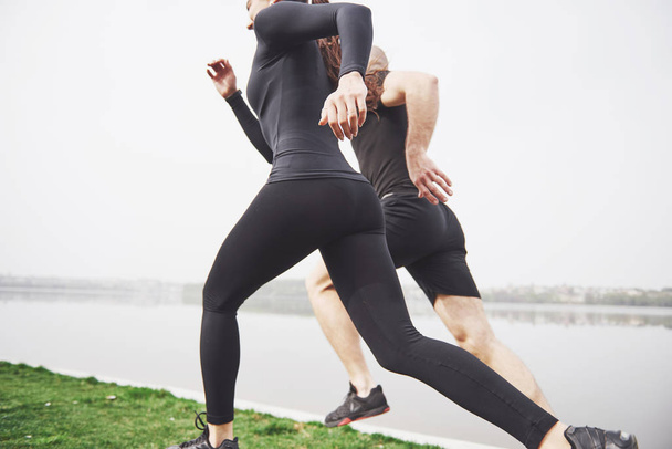 Couple jogging and running outdoors in park near the water. Young bearded man and woman exercising together in morning. - 写真・画像